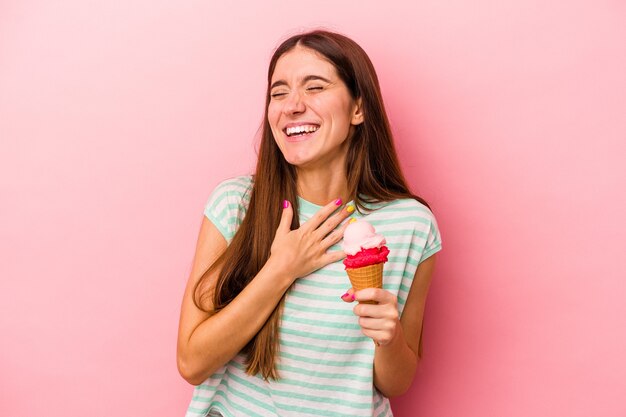 La giovane donna caucasica che tiene un gelato isolato su sfondo rosa ride ad alta voce tenendo la mano sul petto.