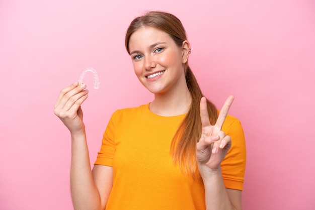 Young caucasian woman holding invisaging isolated on pink background smiling and showing victory sign