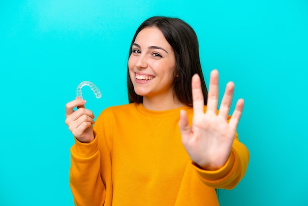 Young caucasian woman holding invisible braces isolated on blue background saluting with hand with happy expression