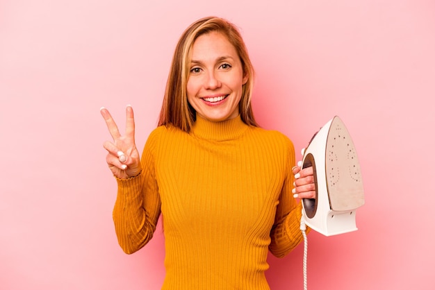 Young caucasian woman holding iron isolated on pink background showing number two with fingers