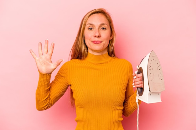 Young caucasian woman holding iron isolated on pink background smiling cheerful showing number five with fingers