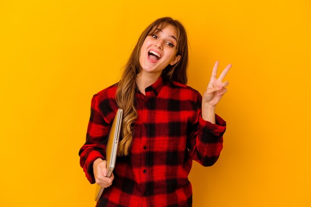 Young caucasian woman holding a laptop isolated joyful and carefree showing a peace symbol with fingers.