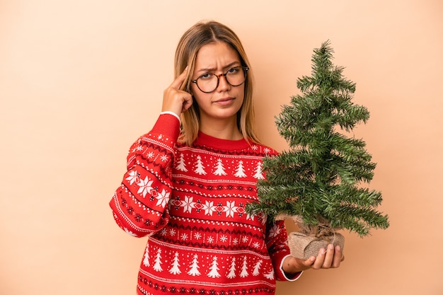 ベージュの背景に分離された小さなクリスマスツリーを指で寺院を指して、考えて、タスクに焦点を当てた若い白人女性。