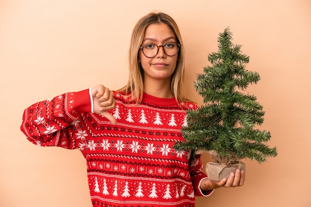 Young caucasian woman holding a little christmas tree isolated on beige background showing a dislike gesture, thumbs down. Disagreement concept.