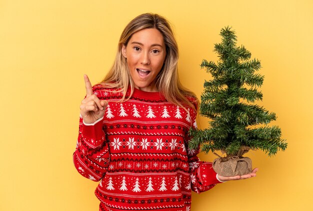 Young caucasian woman holding a little christmas tree isolated on yellow background having an idea, inspiration concept.