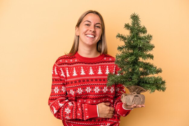 笑って楽しんで黄色の背景に分離された小さなクリスマスツリーを保持している若い白人女性。