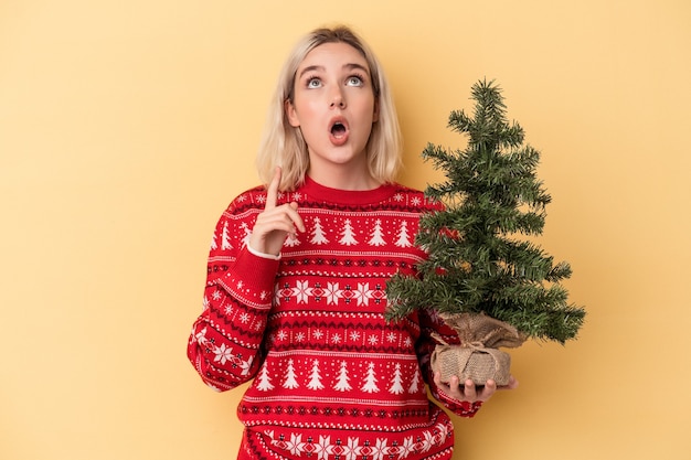 開いた口で逆さまを指している黄色の背景に分離された小さなクリスマスツリーを保持している若い白人女性。