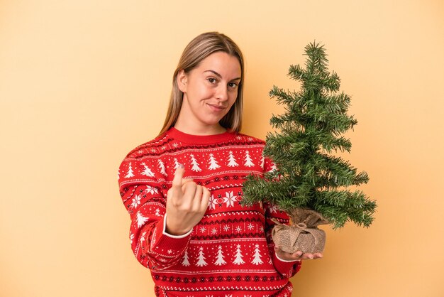 黄色の背景に隔離された小さなクリスマスツリーを持っている若い白人女性は、招待が近づくようにあなたに指を指しています。