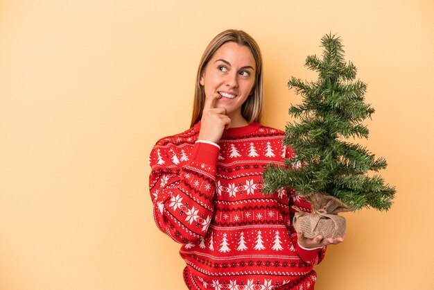 黄色の背景に分離された小さなクリスマスツリーを保持している若い白人女性は、コピースペースを見ている何かについて考えてリラックスしました。