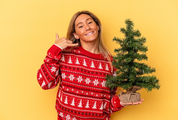 Young caucasian woman holding a little christmas tree isolated on yellow background showing a mobile phone call gesture with fingers.