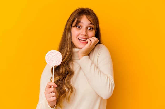 Young caucasian woman holding a lollipop isolated on yellow background biting fingernails, nervous and very anxious.