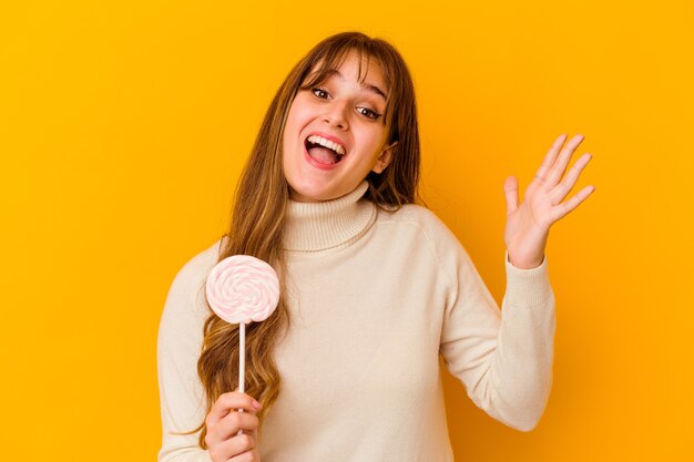 Young caucasian woman holding a lollipop isolated on yellow background receiving a pleasant surprise, excited and raising hands.