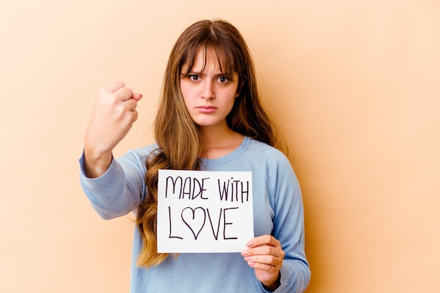 Foto giovane donna caucasica che tiene un fatto con amore cartello isolato mostrando il pugno alla telecamera, aggressiva espressione facciale.