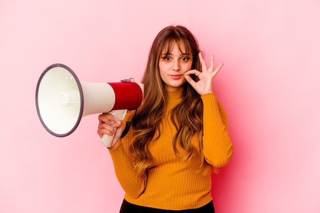 Young caucasian woman holding a megaphone isolated with fingers on lips keeping a secret.