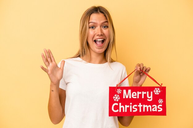 黄色の背景に分離されたメリークリスマスのプラカードを保持している若い白人女性は、嬉しい驚きを受け取り、興奮し、手を上げます。