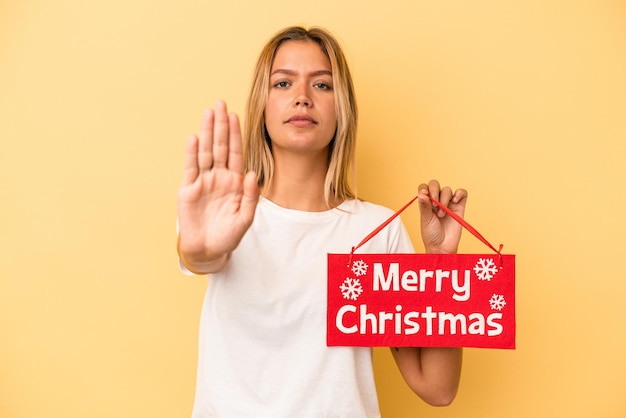 Young caucasian woman holding a merry christmas placard isolated on yellow background standing with outstretched hand showing stop sign, preventing you.