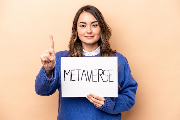 Young caucasian woman holding a metaverse placard isolated on beige background showing number one with finger