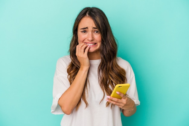 Giovane donna caucasica in possesso di un telefono cellulare isolato su sfondo blu che si morde le unghie, nervosa e molto ansiosa.