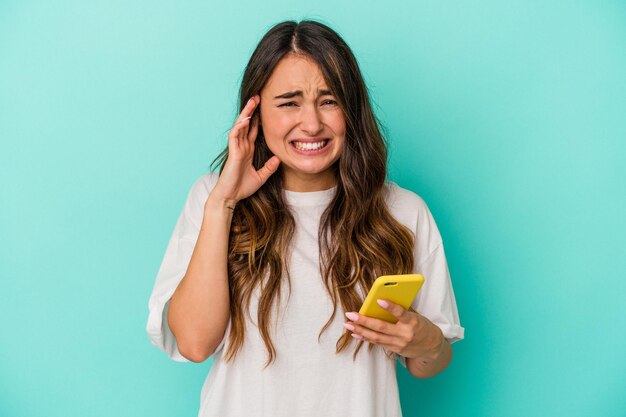 Young caucasian woman holding a mobile phone isolated on blue background covering ears with hands.