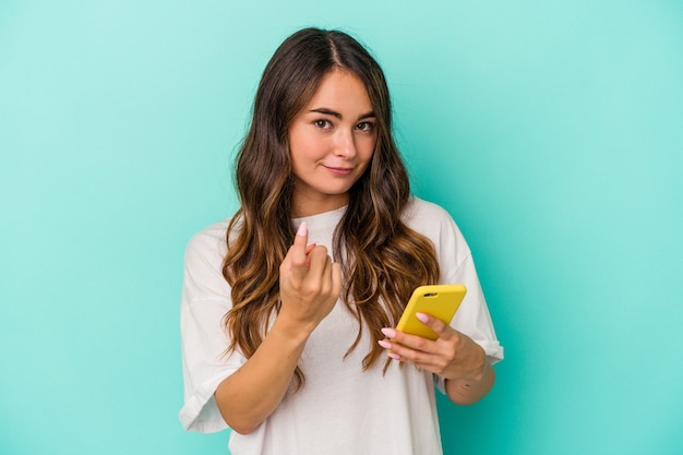 Young caucasian woman holding a mobile phone isolated on blue background pointing with finger at you as if inviting come closer.