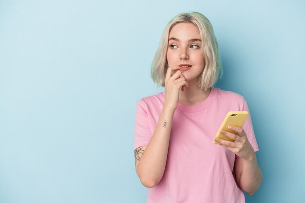 Young caucasian woman holding mobile phone isolated on blue background relaxed thinking about something looking at a copy space.