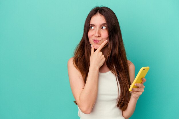 Young caucasian woman holding a mobile phone isolated on blue wall looking sideways with doubtful and skeptical expression.