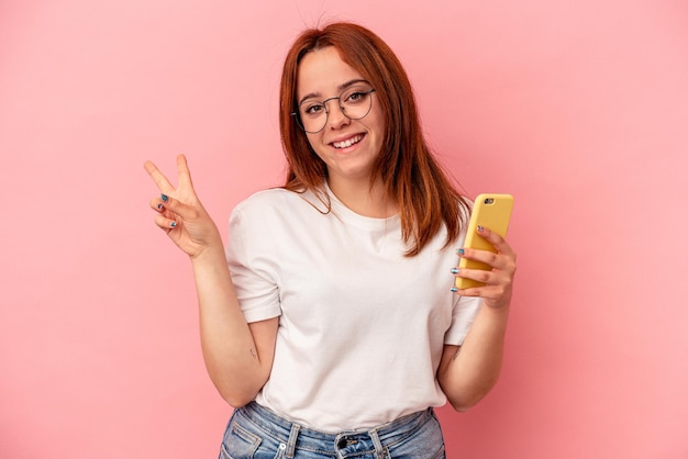 분홍색 배경에 격리된 휴대폰을 들고 있는 젊은 백인 여성은 손가락으로 평화의 상징을 보여주는 즐겁고 평온합니다.