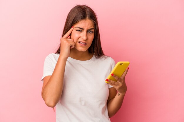 Young caucasian woman holding a mobile phone isolated on pink background showing a disappointment gesture with forefinger.