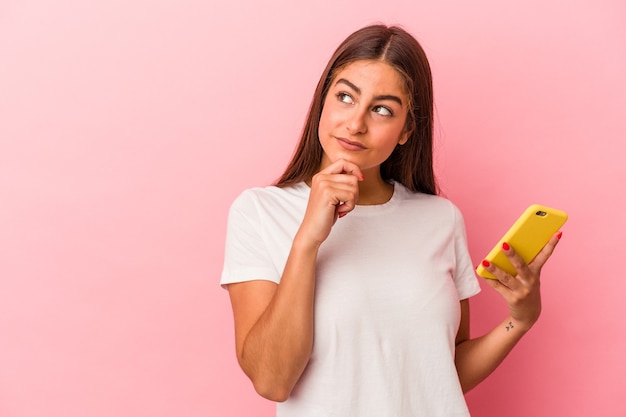 Young caucasian woman holding a mobile phone isolated on pink wall looking sideways with doubtful and skeptical expression.