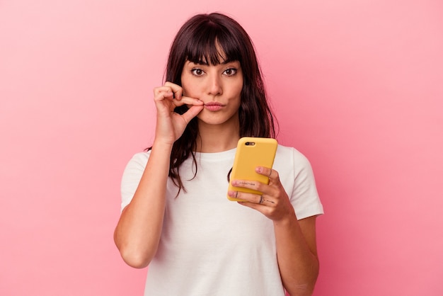 Giovane donna caucasica che tiene un telefono cellulare isolato sulla parete rosa con le dita sulle labbra mantenendo un segreto.