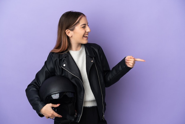 Young caucasian woman holding a motorcycle helmet isolated on purple background pointing finger to the side and presenting a product