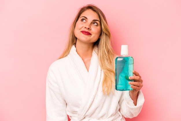 Young caucasian woman holding mouthwash wearing bathrobe isolated on pink background dreaming of achieving goals and purposes