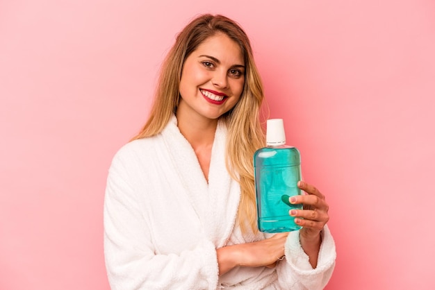 Young caucasian woman holding mouthwash wearing bathrobe isolated on pink background laughing and having fun