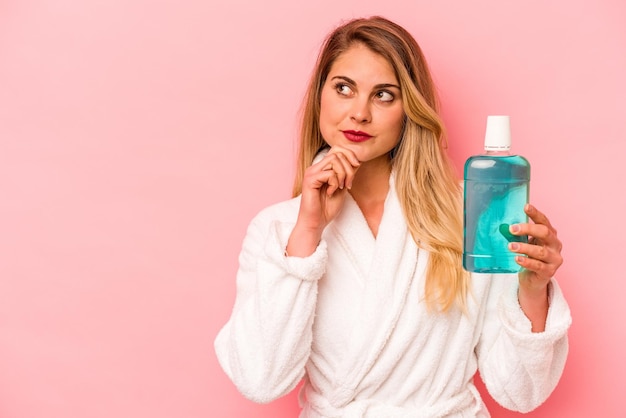 Young caucasian woman holding mouthwash wearing bathrobe isolated on pink background looking sideways with doubtful and skeptical expression