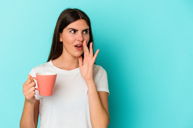 Young caucasian woman holding a mug isolated on blue wall is saying a secret hot braking news and looking aside