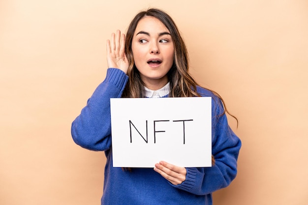 Young caucasian woman holding a nft placard isolated on beige background trying to listening a gossip