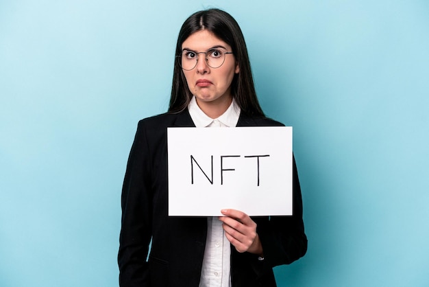 Young caucasian woman holding a NFT placard isolated on blue background shrugs shoulders and open eyes confused