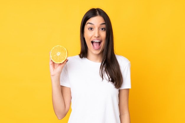 Young caucasian woman holding an orange isolated on yellow wall with surprise and shocked facial expression