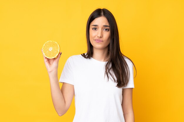 Young caucasian woman holding an orange on yellow wall with sad expression