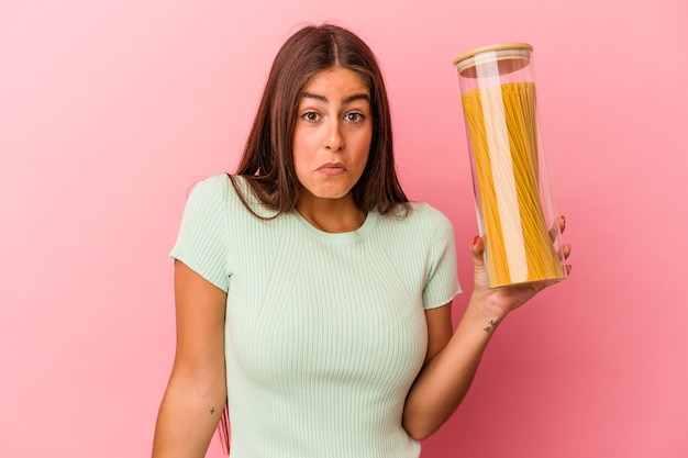 Young caucasian woman holding a pasta jar isolated on pink background shrugs shoulders and open eyes confused.