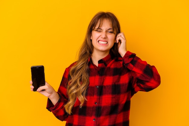 Young caucasian woman holding phone isolated on yellow background covering ears with hands