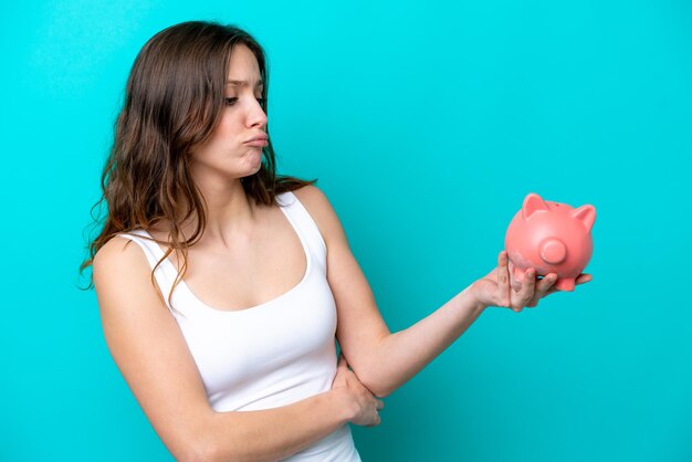 Young caucasian woman holding a piggybank isolated on blue bakcground with sad expression