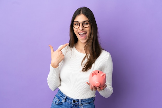 Young caucasian woman holding a piggybank isolated on purple background giving a thumbs up gesture