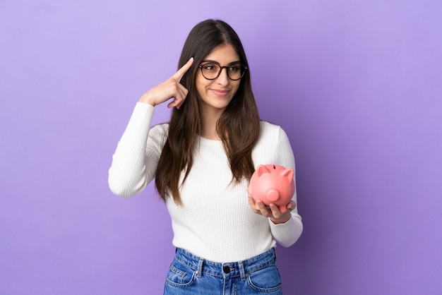 Young caucasian woman holding a piggybank isolated on purple background having doubts and thinking