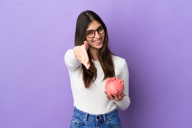 Young caucasian woman holding a piggybank isolated on purple background shaking hands for closing a good deal