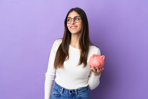 Young caucasian woman holding a piggybank isolated on purple background thinking an idea while looking up