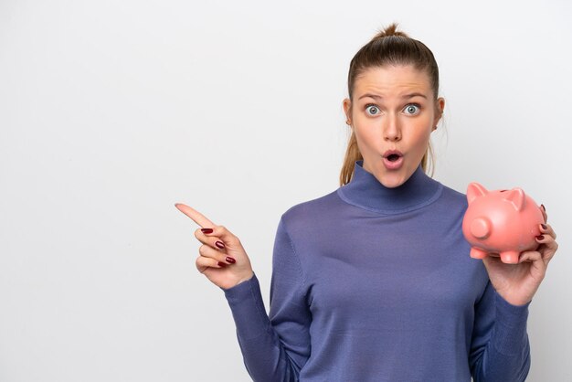 Young caucasian woman holding a piggybank isolated on white background surprised and pointing side