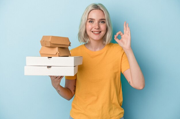 Young caucasian woman holding pizzas and burgers isolated on blue background cheerful and confident showing ok gesture.