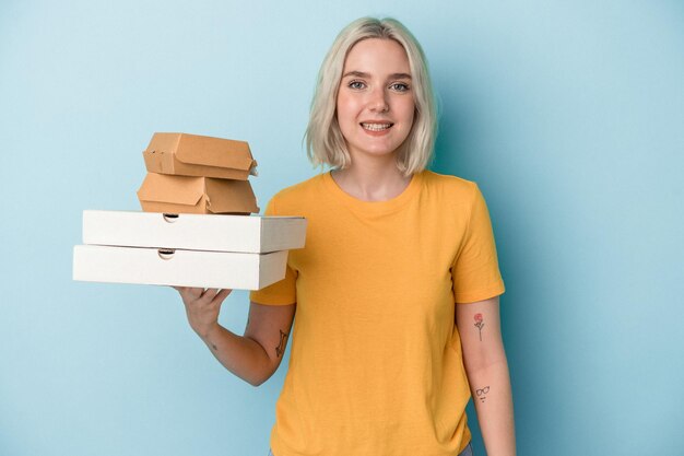 Молодая кавказская женщина, держащая пиццу и гамбургеры на синем фоне, счастливая, улыбающаяся и веселая