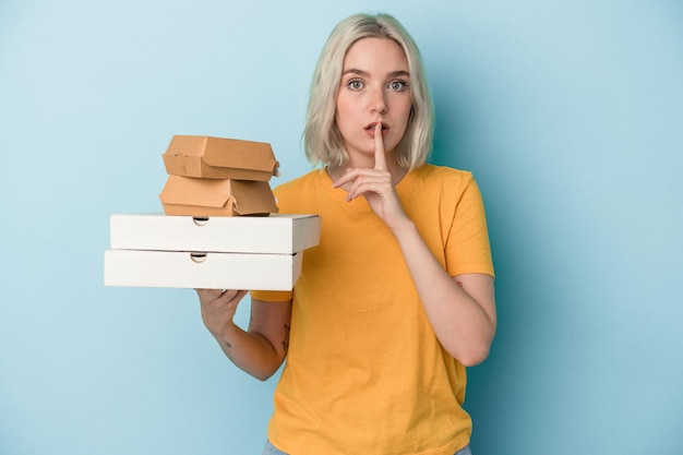 Photo young caucasian woman holding pizzas and burgers isolated on blue background keeping a secret or asking for silence